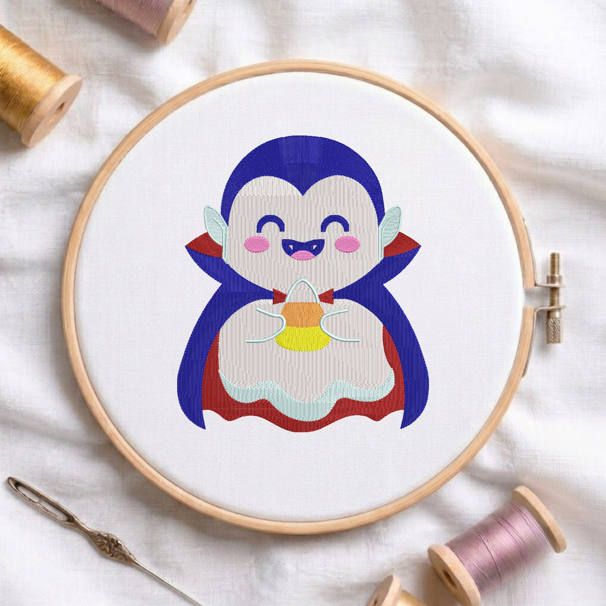 A circular embroidery hoop holding white fabric with a cartoon vampire penguin design in blue, red, yellow, pink, and white. The hoop is photographed on a white fabric surface with spools of thread and a needle around it.