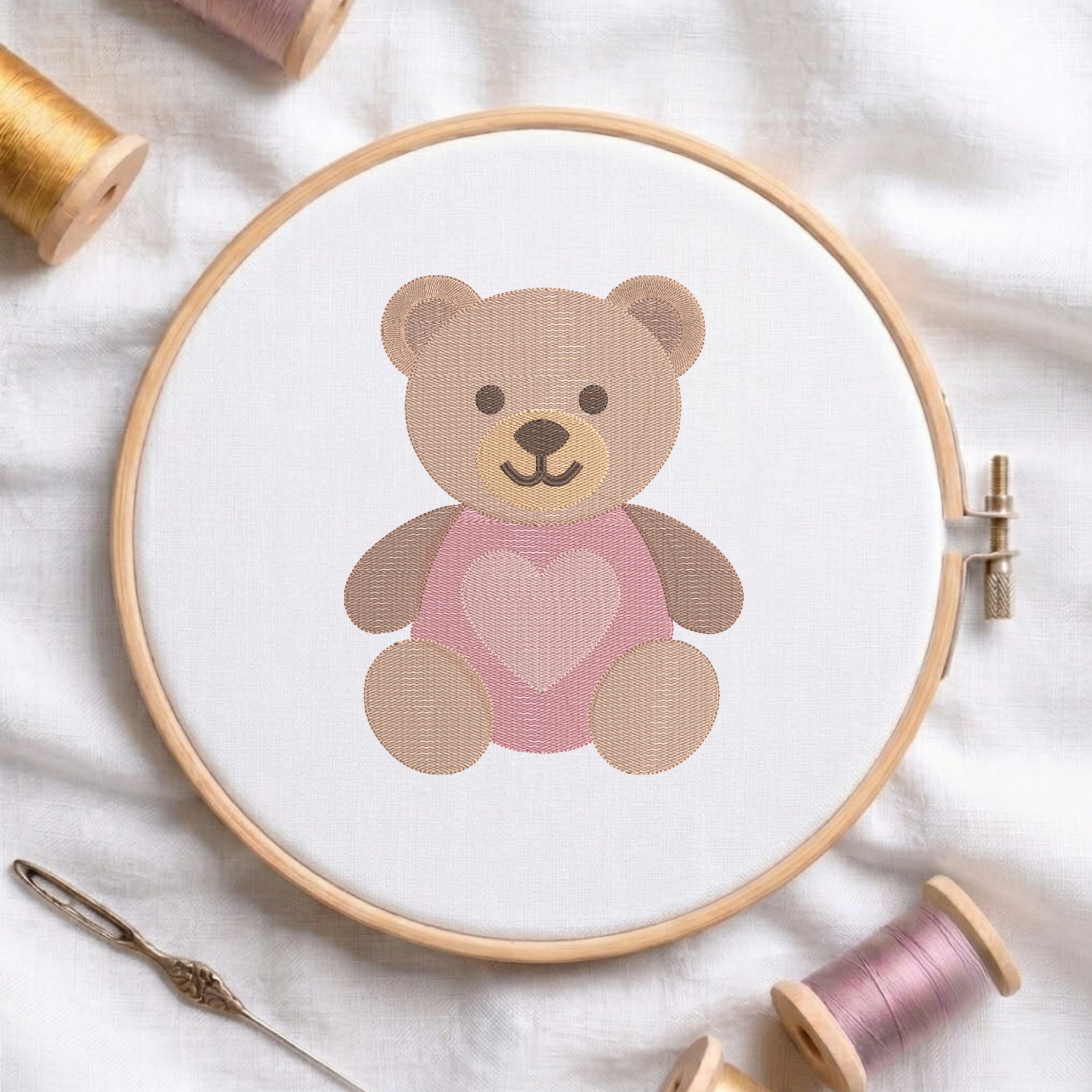 A wooden embroidery hoop holds white fabric with a teddy bear design wearing a pink heart-patterned sweater. The hoop is styled on a white fabric background with spools of thread and a needle nearby.