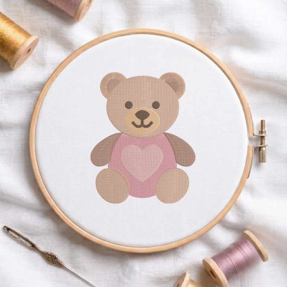 A wooden embroidery hoop holds white fabric with a teddy bear design wearing a pink heart-patterned sweater. The hoop is styled on a white fabric background with spools of thread and a needle nearby.