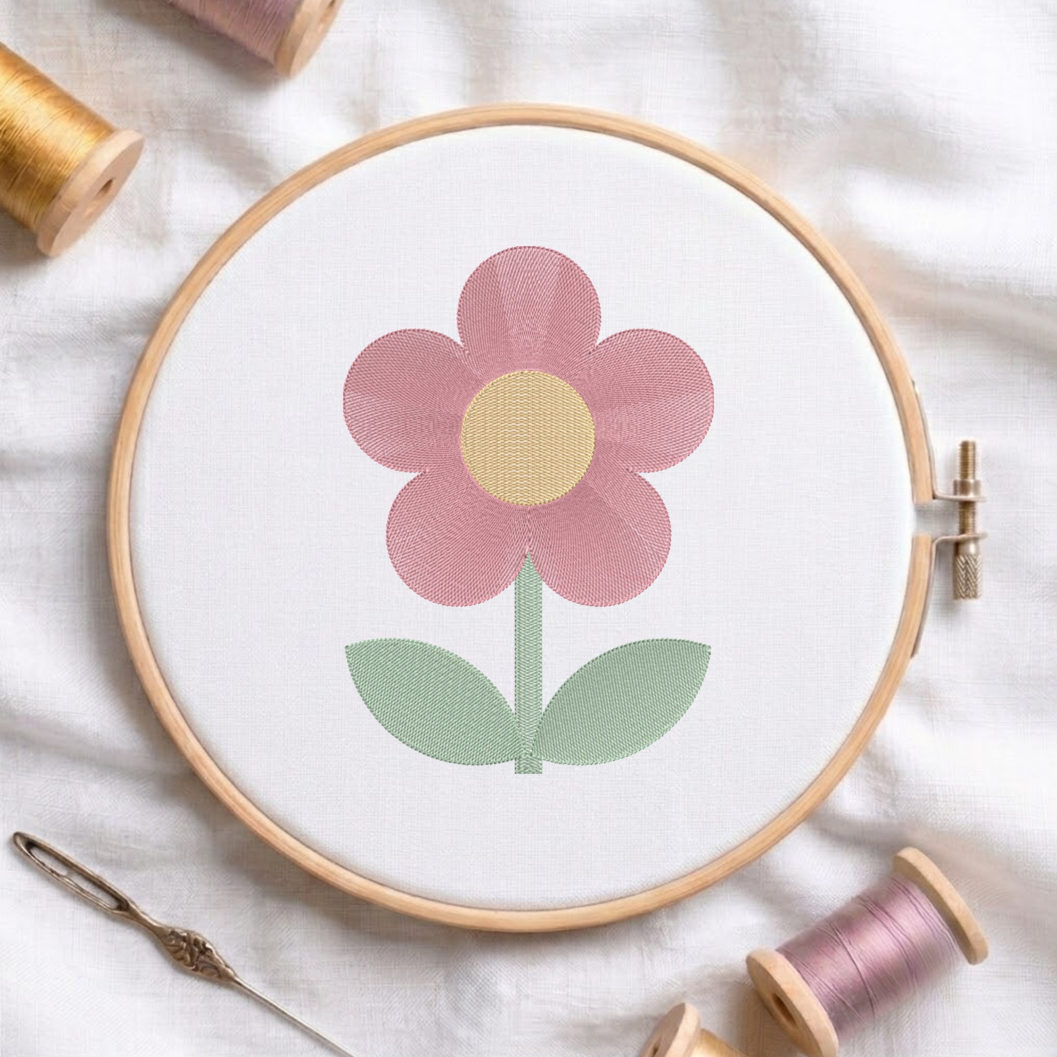 A wooden embroidery hoop holds white fabric with a stylized flower design in pink, yellow, and green. Surrounding props include spools of thread, a needle, and soft white fabric.