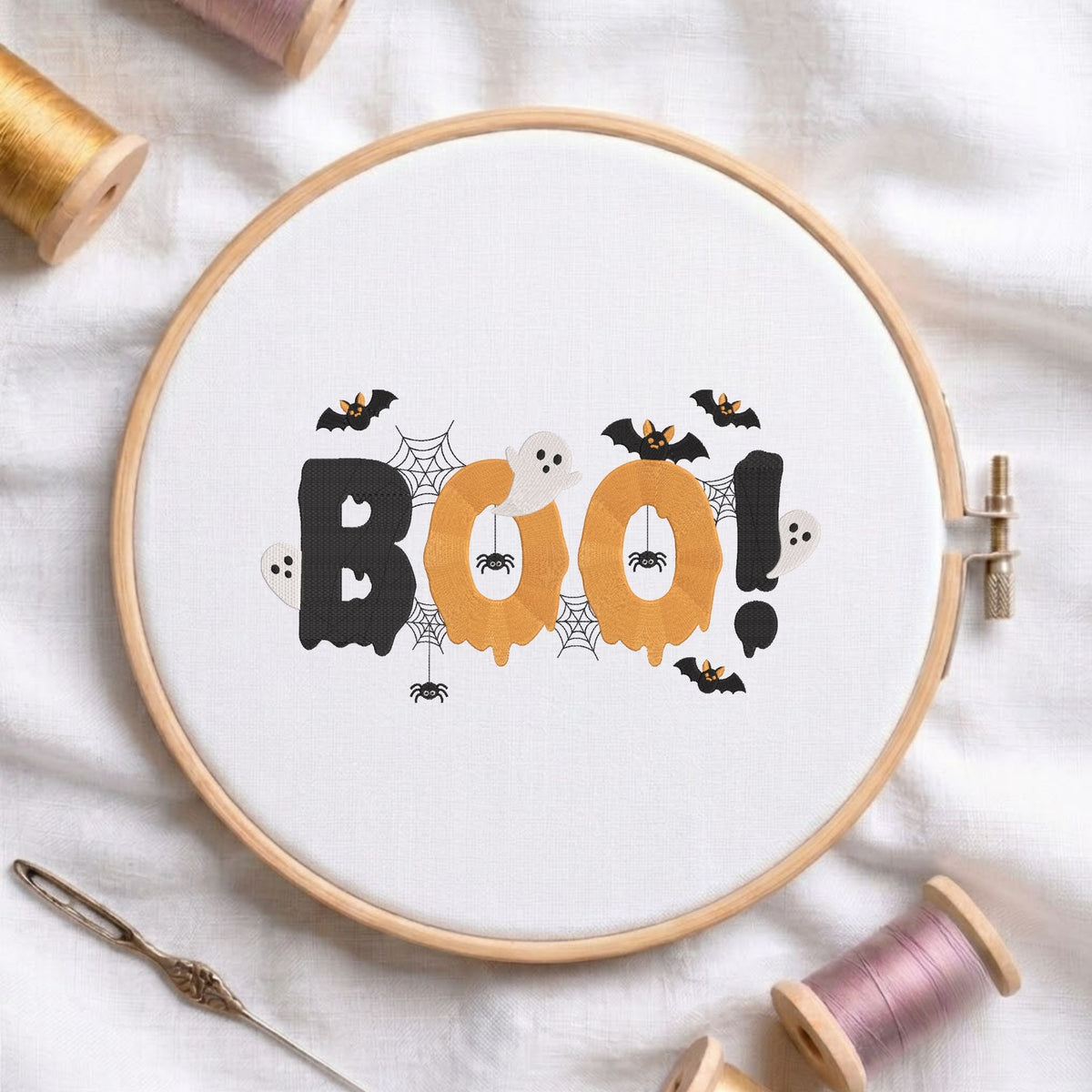 Spooky "BOO!" lettering with cute ghosts, bats, spiders, and webs machine embroidery design displayed in a wooden hoop on white fabric with thread spools and a needle.   