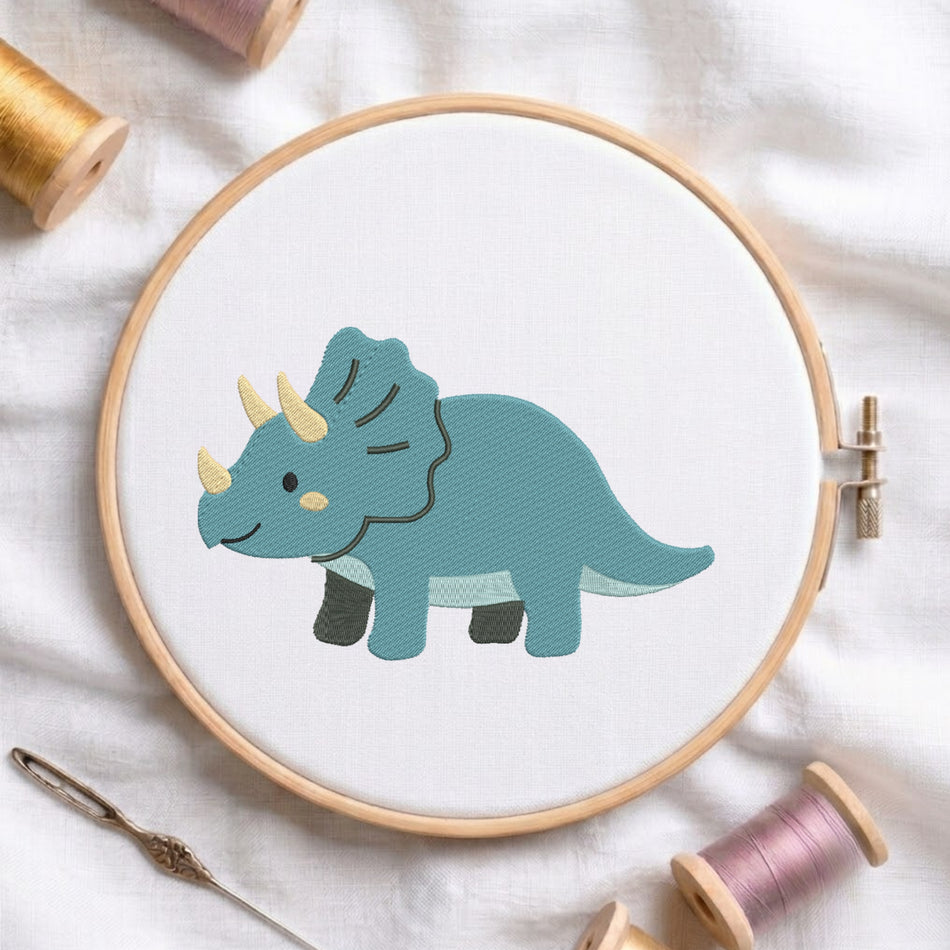 A wooden embroidery hoop holds white fabric with a stitched cartoon triceratops dinosaur in teal, black, and pale yellow. The hoop is styled on a white fabric background with spools of thread and a needle around it.