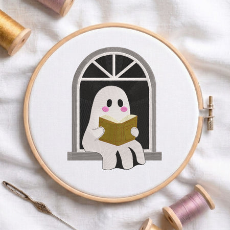 A circular embroidery hoop on white fabric holds a stitched design of a white ghost reading a gold book in front of a gray arched window. A needle lies at the lower left, and spools of thread are partially visible around the hoop.