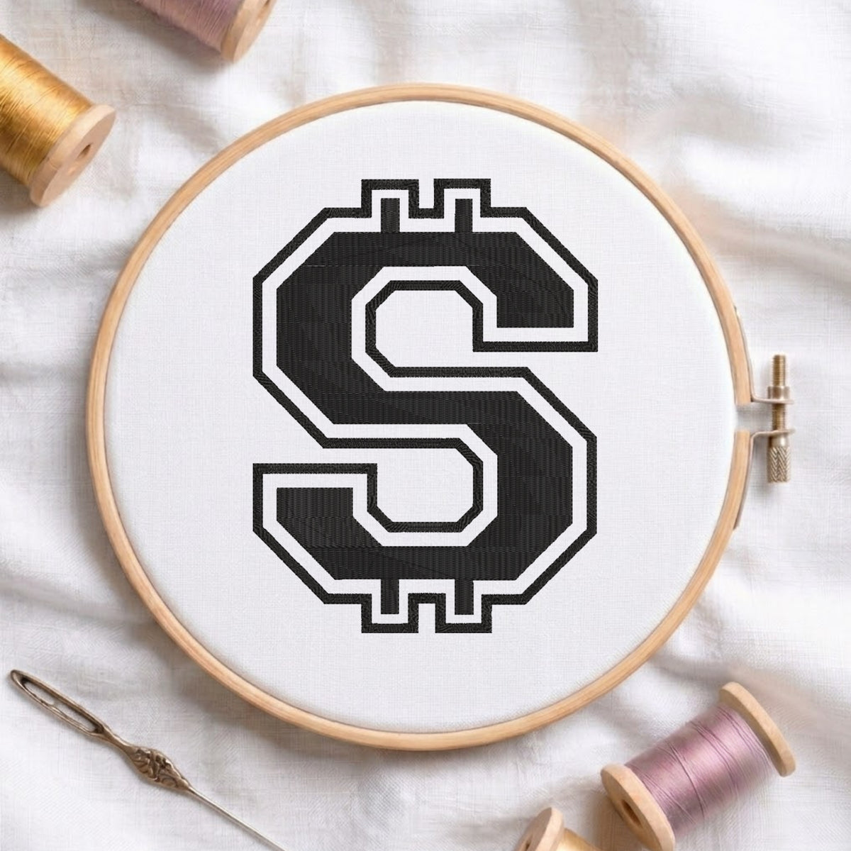 A circular embroidery hoop holding white fabric with a large black-and-white stylized letter S motif, photographed on a white cloth with spools of thread and a needle around it.