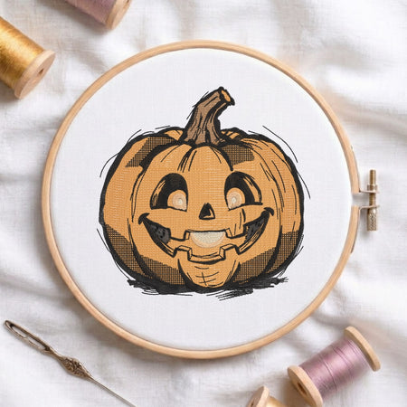 A circular embroidery hoop holding white fabric with a printed or stitched jack-o'-lantern pumpkin design in orange, black, and brown. The hoop is photographed on a white fabric surface with spools of thread and a needle around it.