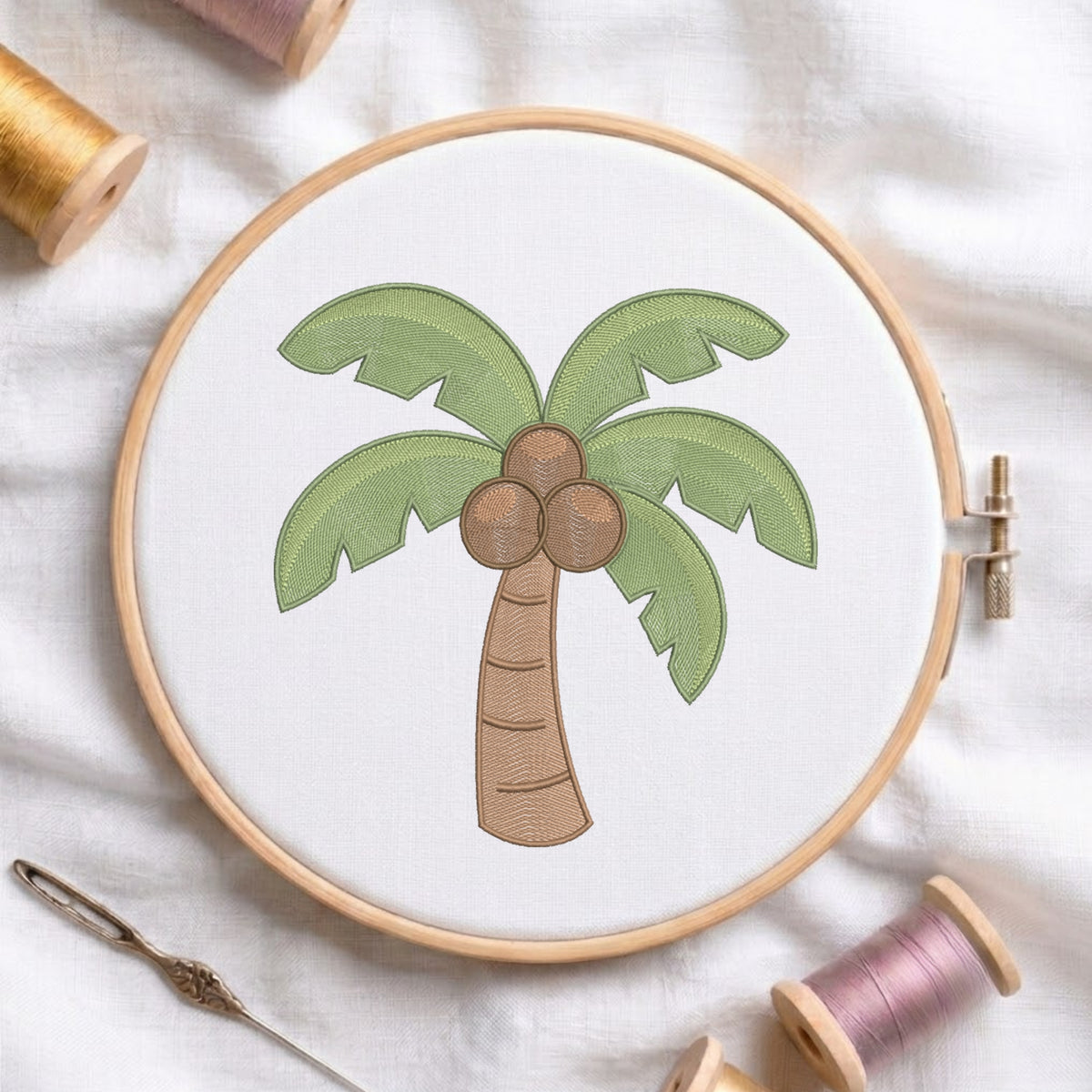 A circular embroidery hoop holding white fabric with a palm tree motif in green, brown, and beige tones, styled on a white fabric background with spools of thread and a needle nearby.
