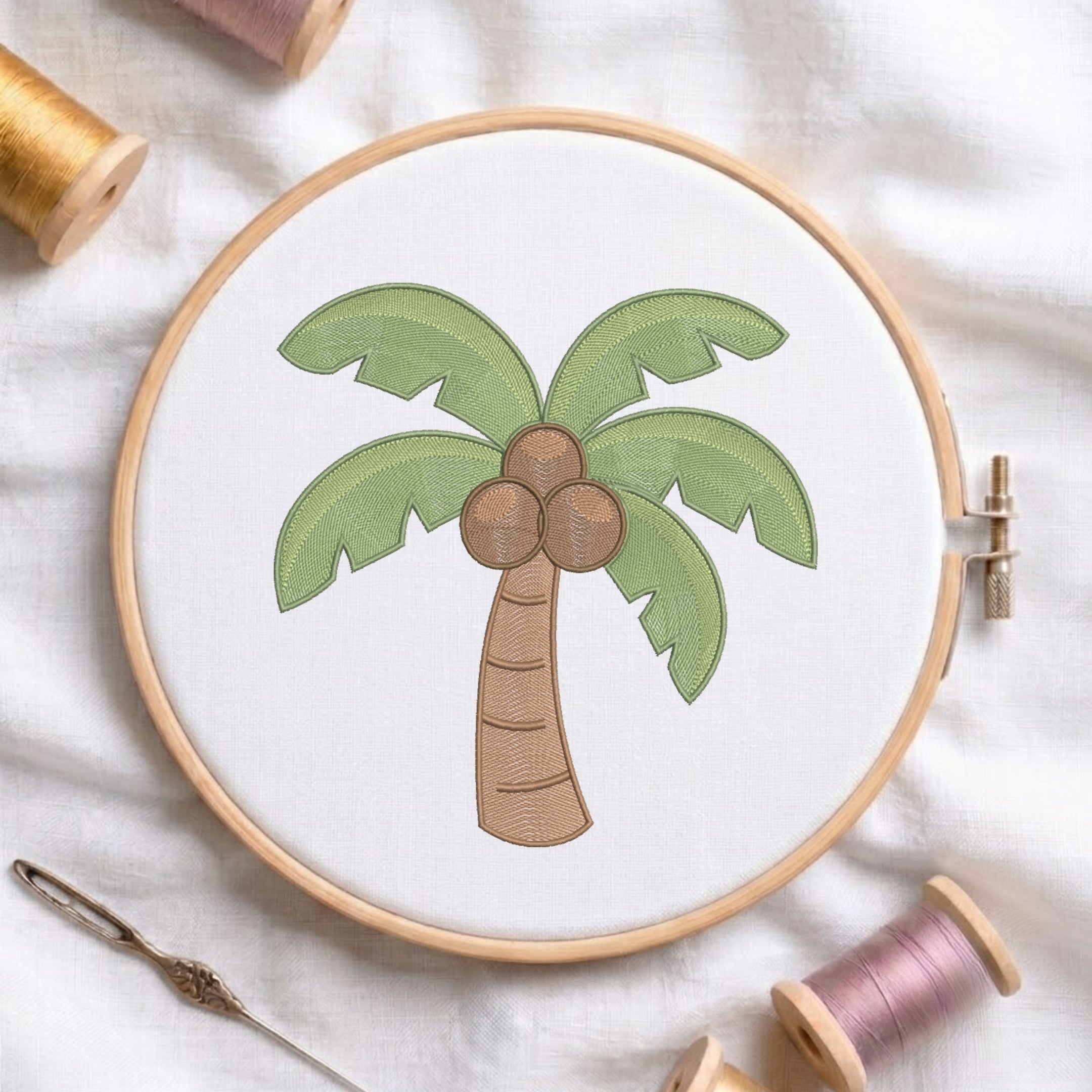 A circular embroidery hoop holding white fabric with a palm tree motif in green, brown, and beige tones, styled on a white fabric background with spools of thread and a needle nearby.