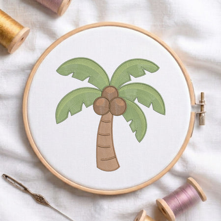 A circular embroidery hoop holding white fabric with a palm tree motif in green, brown, and beige tones, styled on a white fabric background with spools of thread and a needle nearby.