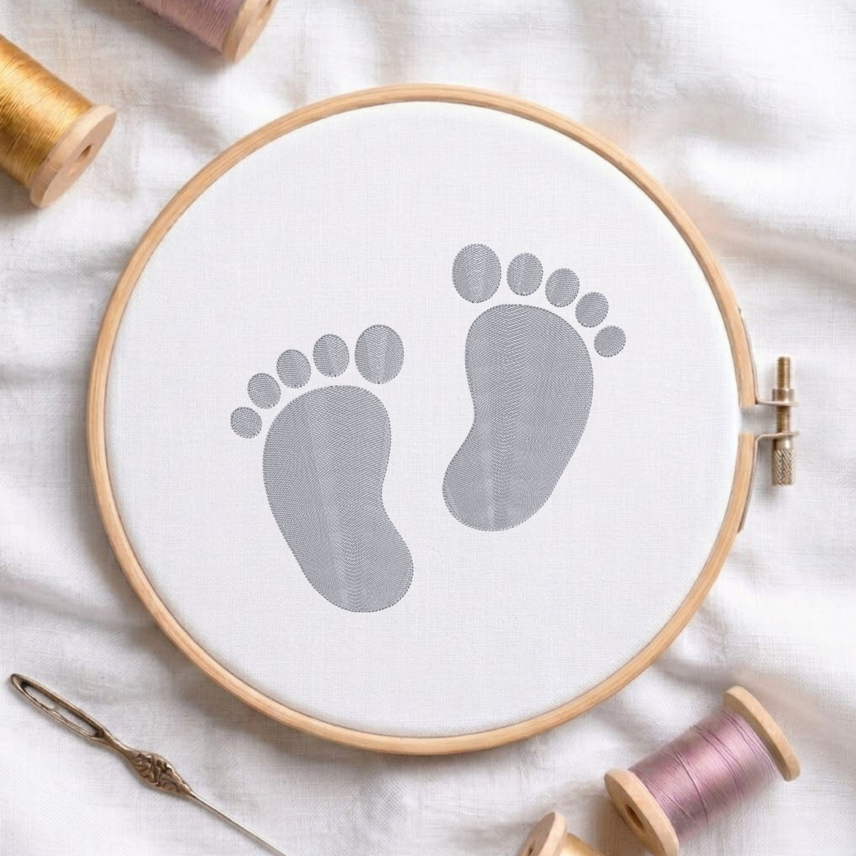 A circular embroidery hoop holding white fabric with a gray baby footprint design, styled on a white cloth with spools of thread and a needle around it.