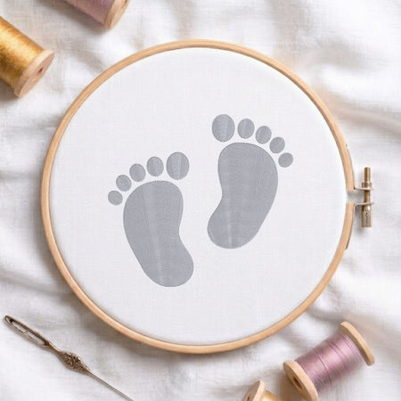 A circular embroidery hoop holding white fabric with a gray baby footprint design, styled on a white cloth with spools of thread and a needle around it.