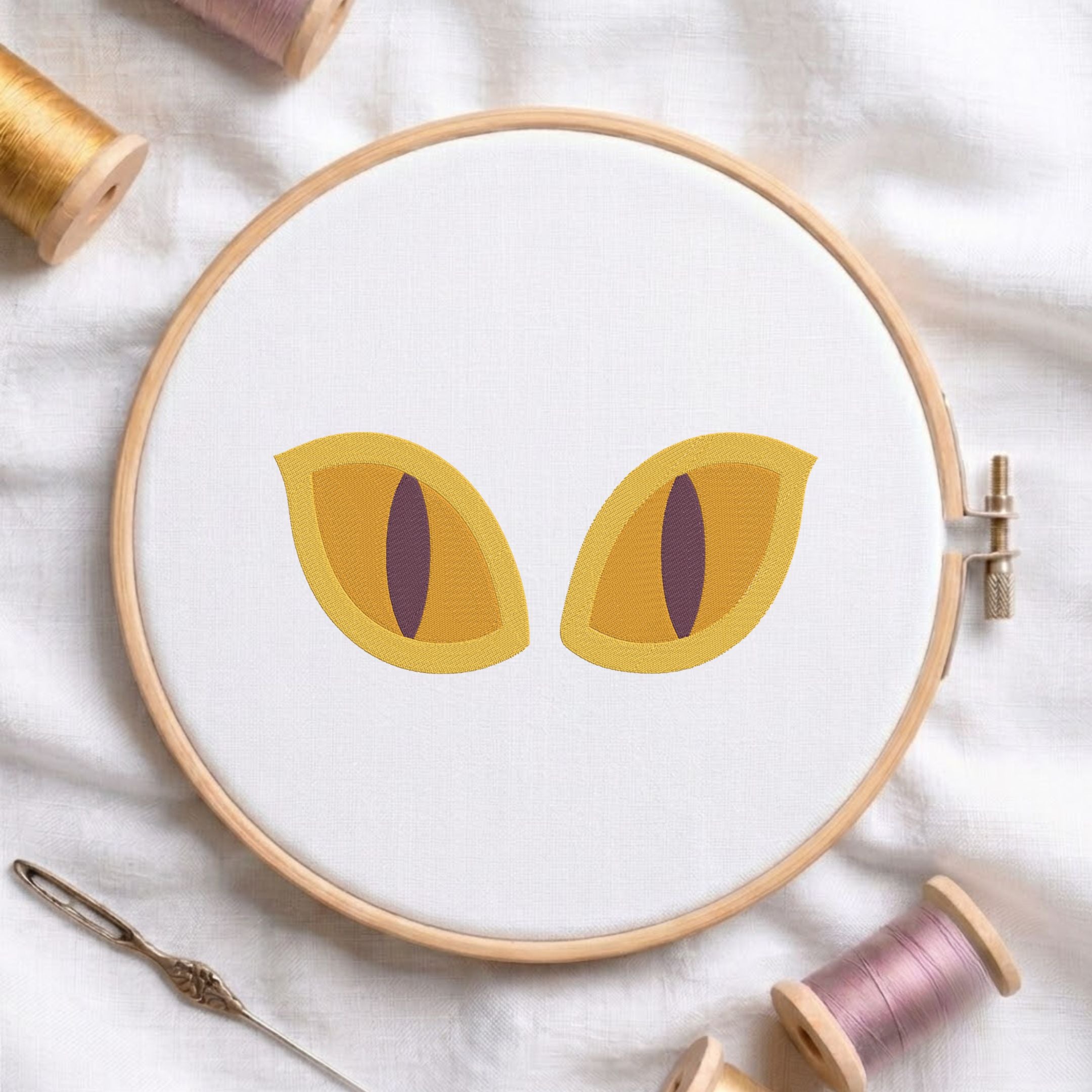 A circular embroidery hoop on white fabric, featuring two stylized yellow cat eyes with dark purple slit pupils. Spools of thread and a needle are arranged around the hoop on a soft white fabric background.