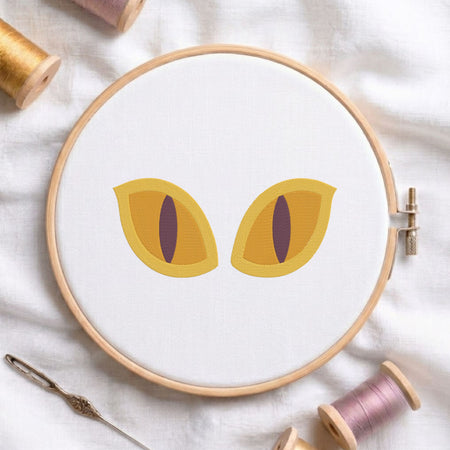 A circular embroidery hoop on white fabric, featuring two stylized yellow cat eyes with dark purple slit pupils. Spools of thread and a needle are arranged around the hoop on a soft white fabric background.
