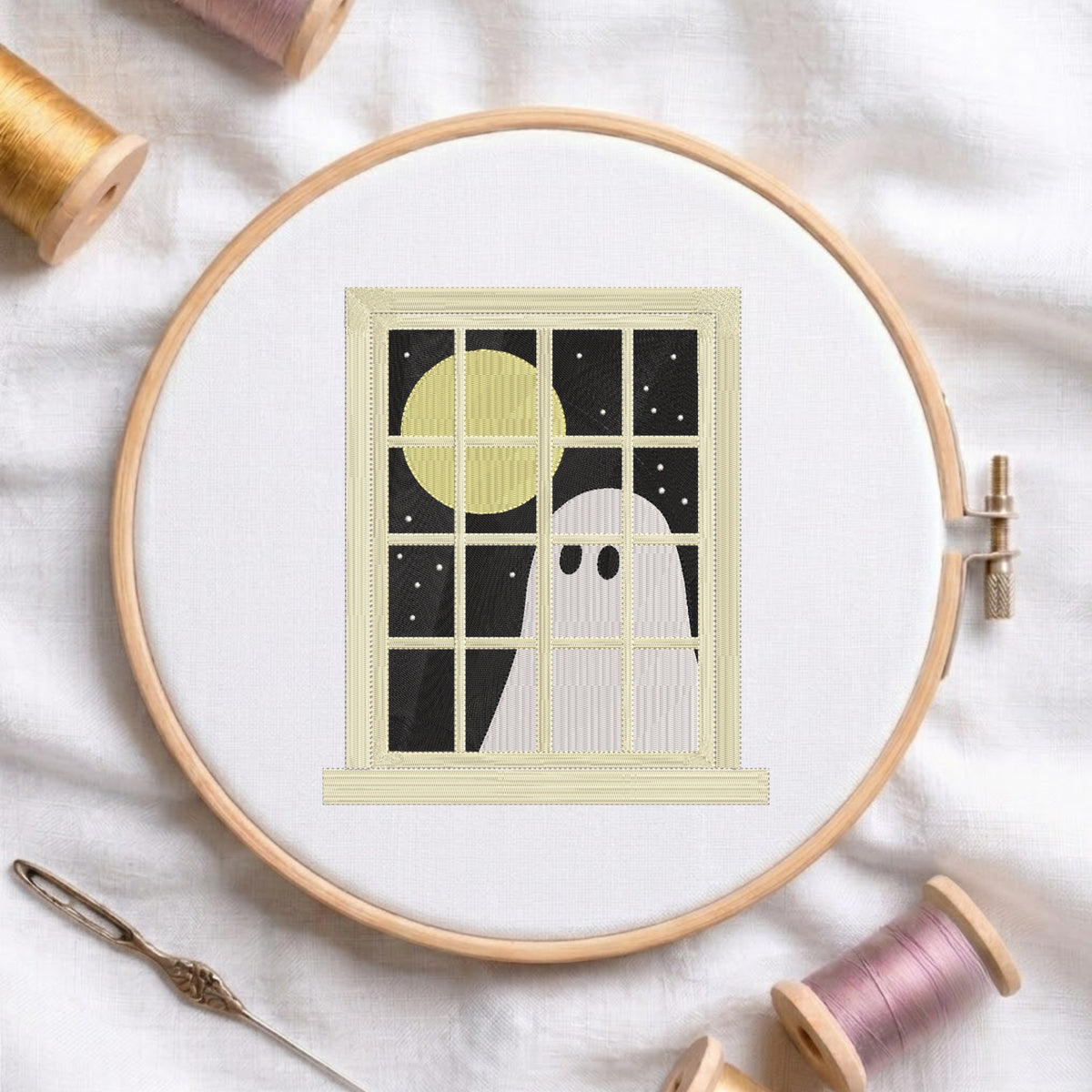 A wooden embroidery hoop holds a white fabric with a stitched design of a ghost looking out a window at a yellow moon and black night sky. A metal needle lies nearby on white fabric, with spools of thread around the hoop.