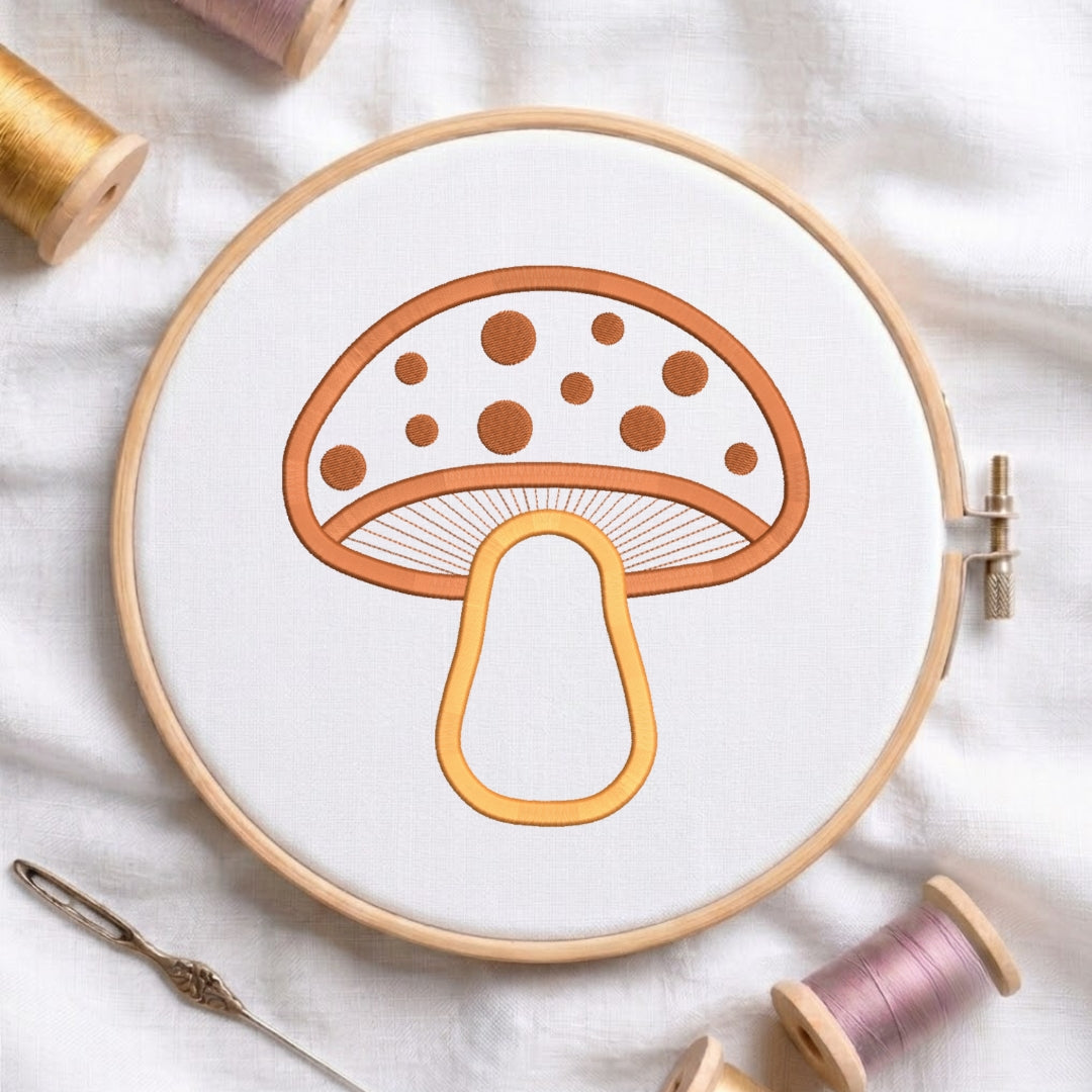 A mushroom embroidery design held in a wooden hoop on white fabric, surrounded by spools of thread and a needle.