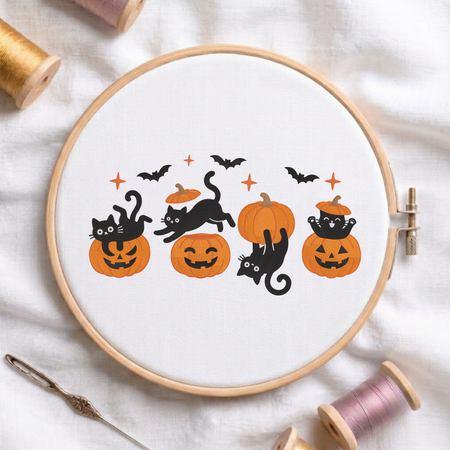 Playful black cats acrobatically posing on carved jack-o-lantern pumpkins with flying bats Halloween machine embroidery design shown in a wooden embroidery hoop on white fabric with thread spools and a needle