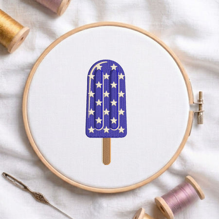 Purple ice cream popsicle with white star patterns and a wooden stick machine embroidery design, displayed inside a round wooden hoop on white fabric with thread spools and a needle.