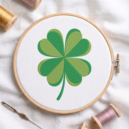 A wooden embroidery hoop holds white fabric with a green shamrock design stitched in the center. A needle and thread spools are arranged around it on a white fabric background.