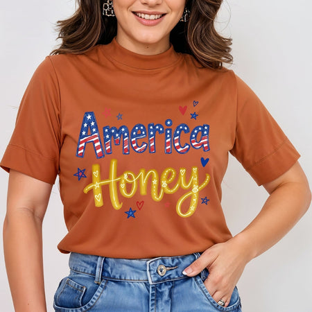 A woman wearing a rust-orange mock-neck t-shirt with a patriotic graphic that reads 'America Honey' in red, white, and blue lettering with yellow glitter-style script, paired with blue jeans.