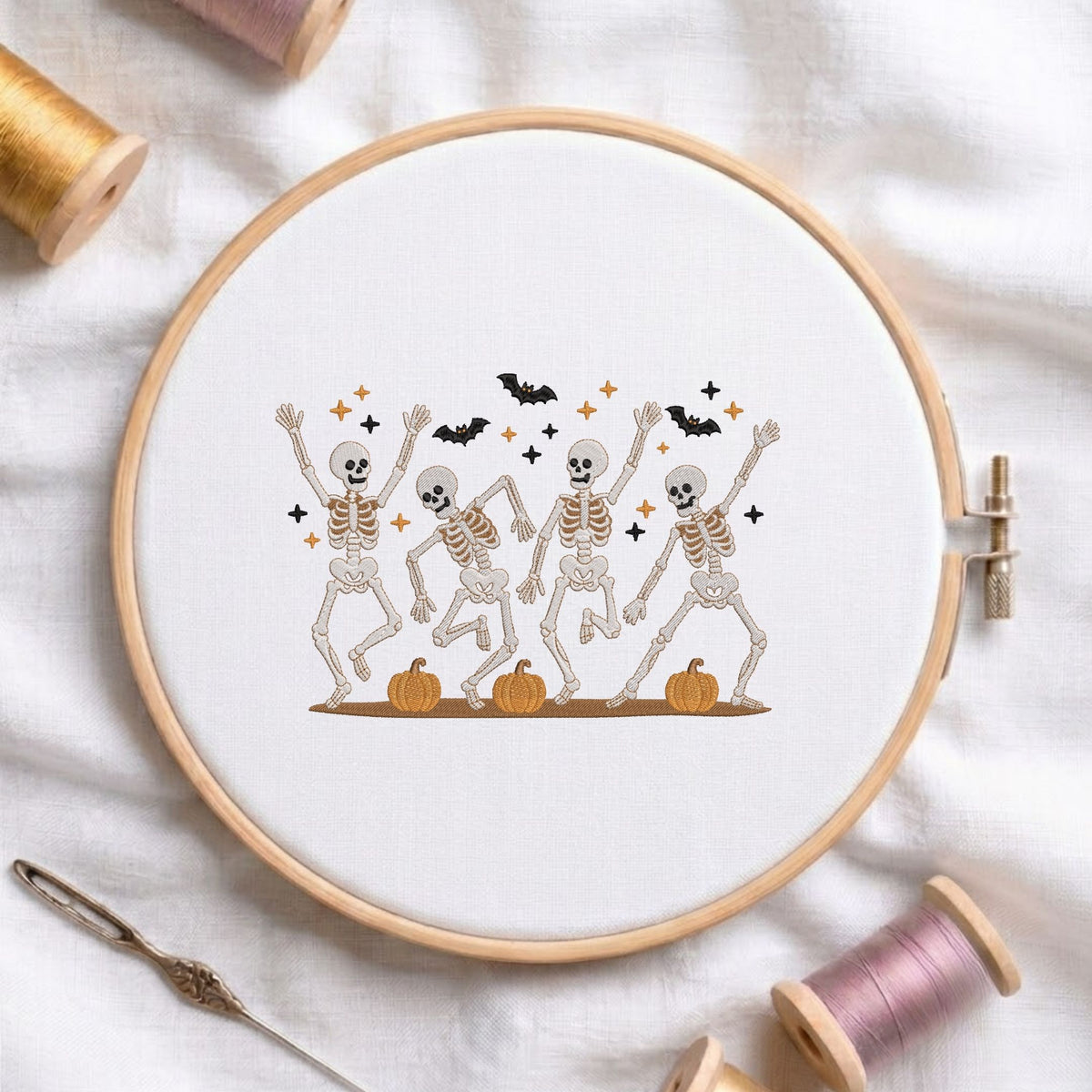 Cute dancing skeletons with pumpkins and bats Halloween machine embroidery design displayed in a wooden hoop on white fabric with thread spools and a needle   