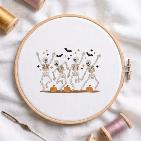 Cute dancing skeletons with pumpkins and bats Halloween machine embroidery design displayed in a wooden hoop on white fabric with thread spools and a needle   
