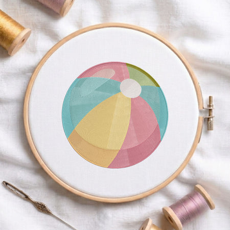 A circular embroidery hoop holding white fabric with a colorful beach ball design stitched in pastel pink, teal, yellow, and green. The hoop is styled on a white fabric background with spools of thread and a needle.