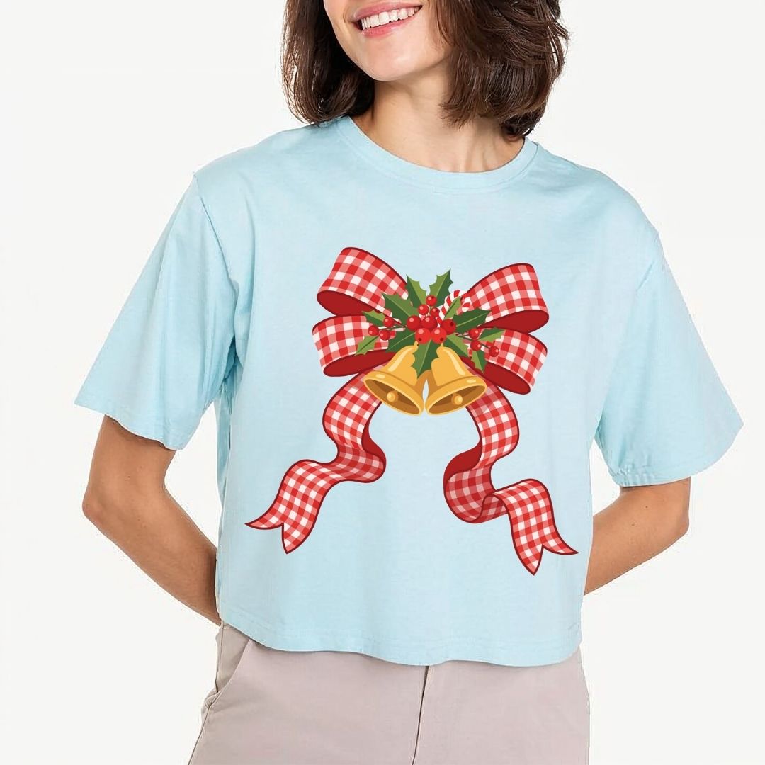 A woman wearing a light blue cropped T-shirt with a festive Christmas graphic of two gold bells, a red and white gingham ribbon, and green holly leaves, paired with light beige pants against a plain light background.