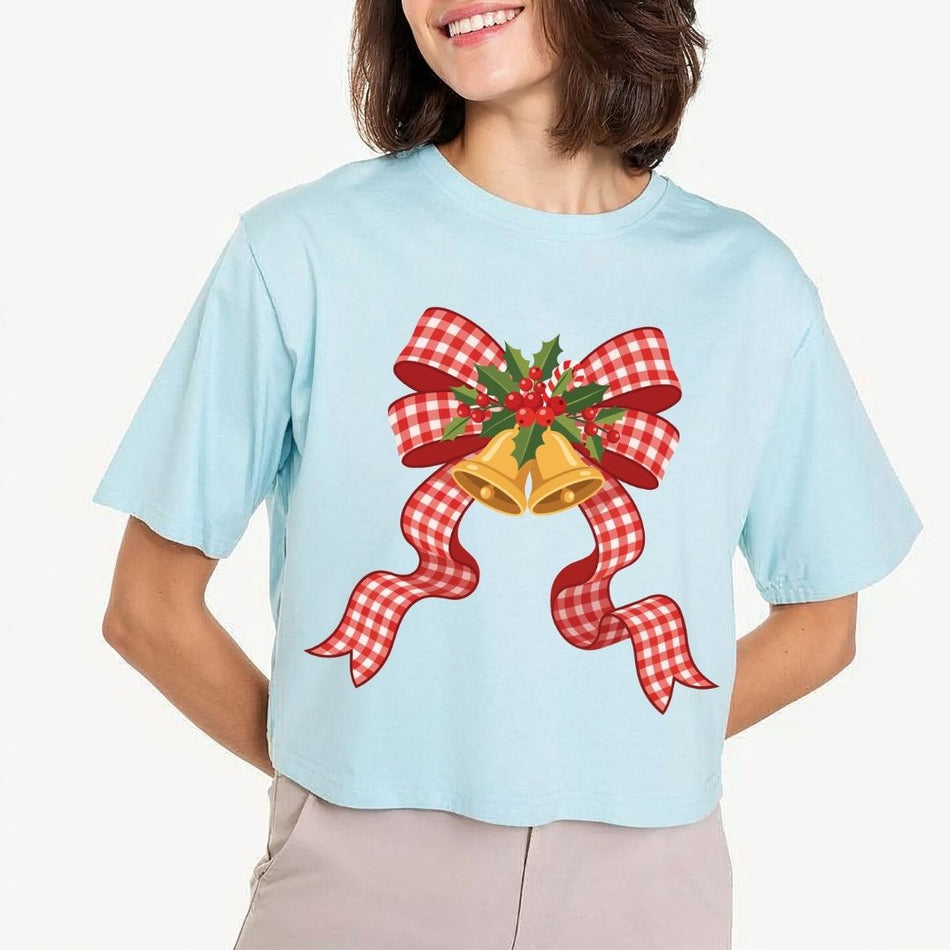 A woman wearing a light blue cropped T-shirt with a festive Christmas graphic of two gold bells, a red and white gingham ribbon, and green holly leaves, paired with light beige pants against a plain light background.