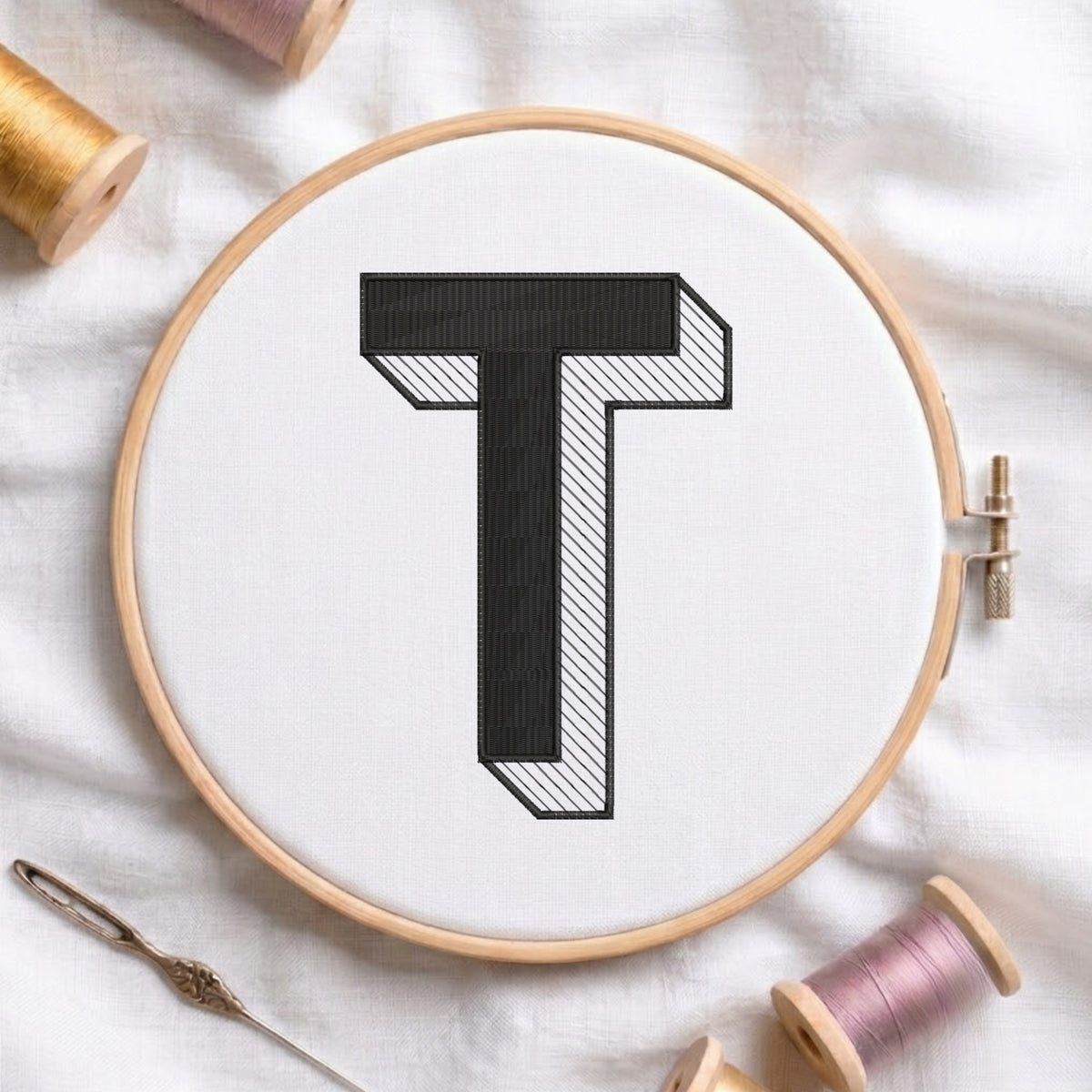An embroidered 3D block letter "T" with striped shading texture, stitched inside a wooden hoop on white fabric with sewing threads.