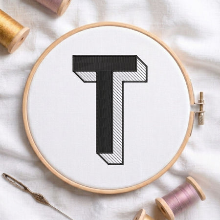 An embroidered 3D block letter "T" with striped shading texture, stitched inside a wooden hoop on white fabric with sewing threads.
