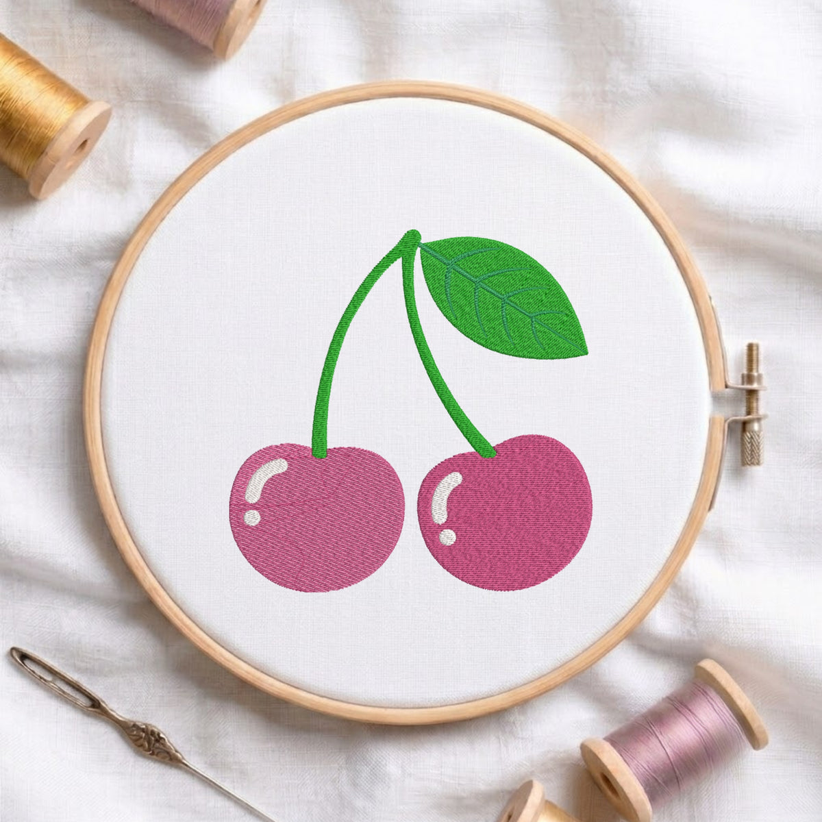 A cherry embroidery design held in a wooden embroidery hoop, surrounded by spools of thread and a needle on a white fabric background.