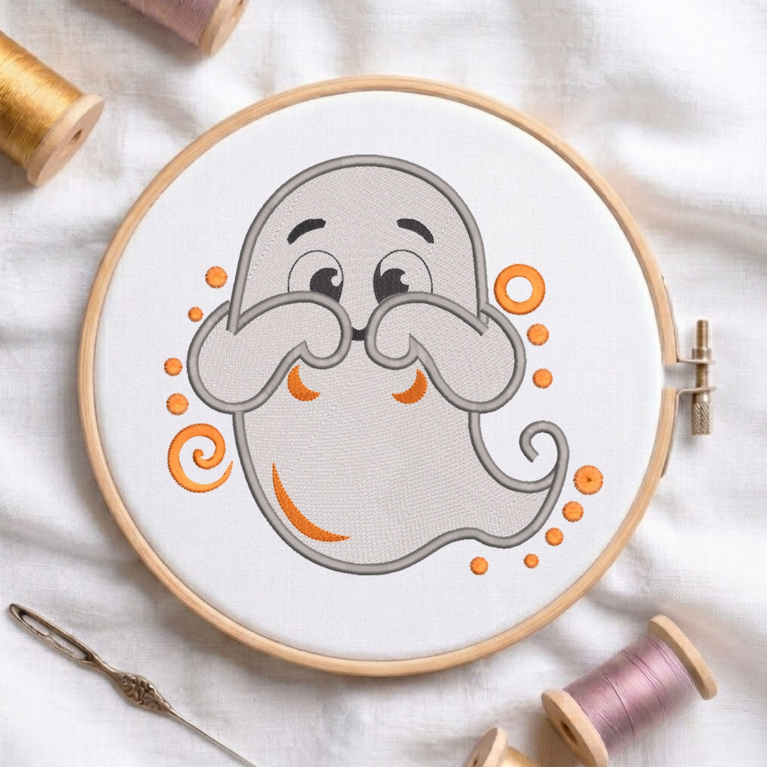 A circular embroidery hoop holding white fabric with a cartoon ghost design in gray and orange, surrounded by spools of thread and a needle on a white fabric background.