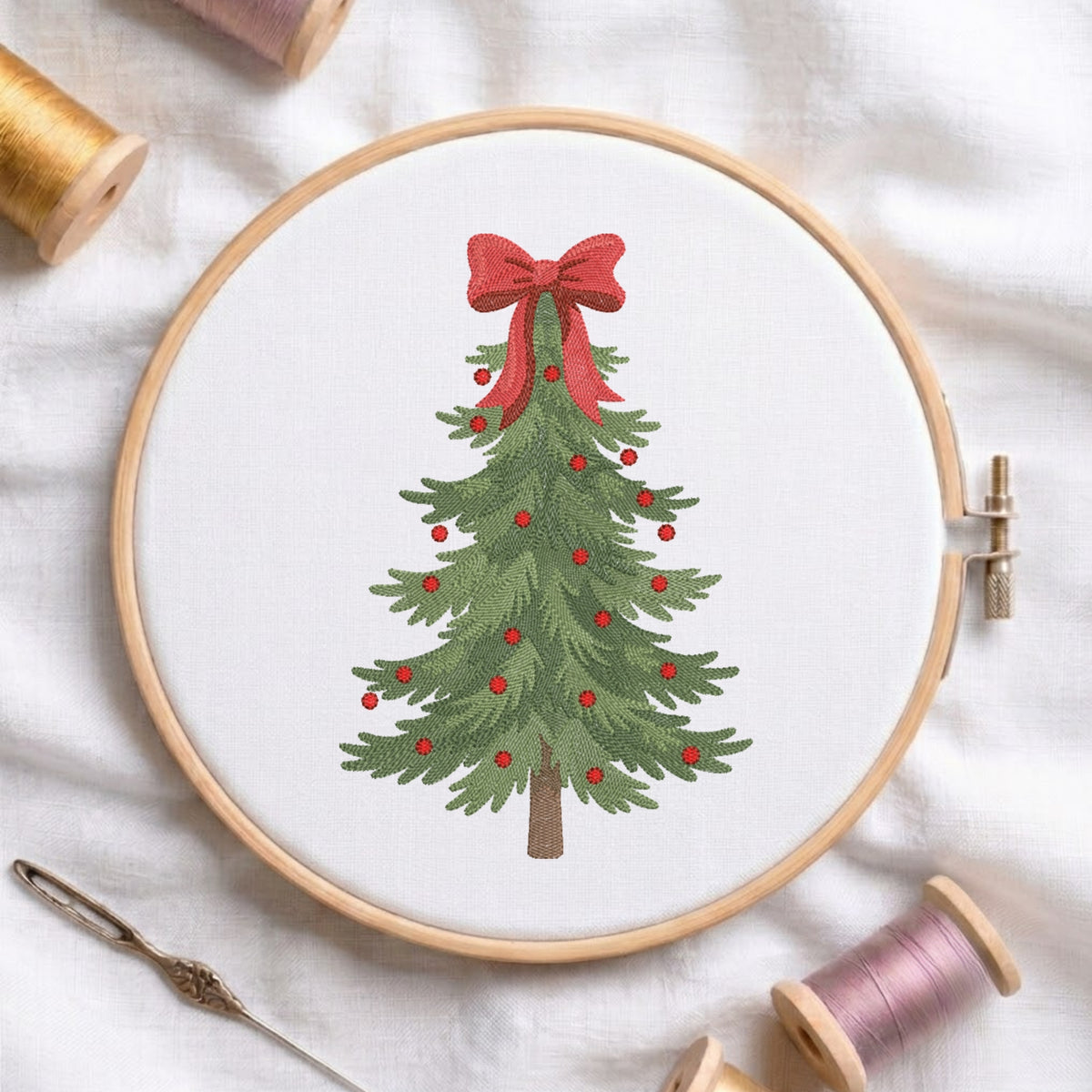A circular embroidery hoop holds white fabric with a green Christmas tree design decorated with small red berries and a large red bow at the top. The hoop is photographed on a white fabric surface with spools of thread and a needle around it.