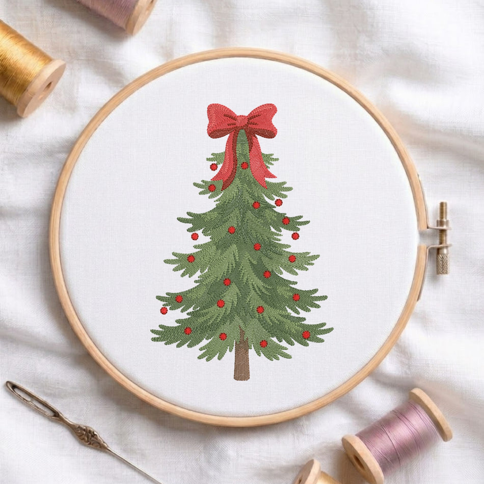 A circular embroidery hoop holds white fabric with a green Christmas tree design decorated with small red berries and a large red bow at the top. The hoop is photographed on a white fabric surface with spools of thread and a needle around it.