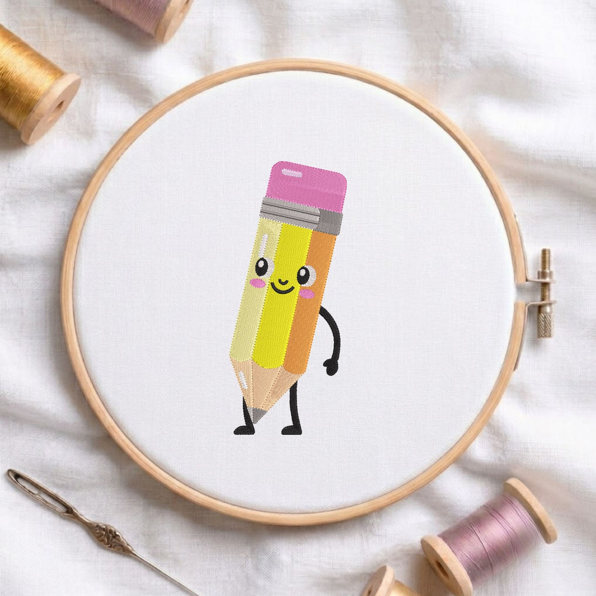 Cute cartoon pencil character with a smiling face and pink cheeks machine embroidery design, displayed inside a round wooden hoop on white fabric with thread spools and a needle.