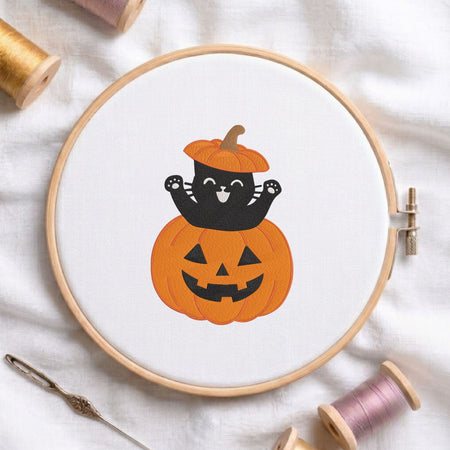 Cute black cat wearing a pumpkin hat sitting inside a jack-o-lantern Halloween machine embroidery design shown in a wooden embroidery hoop on white fabric with thread spools and a needle
