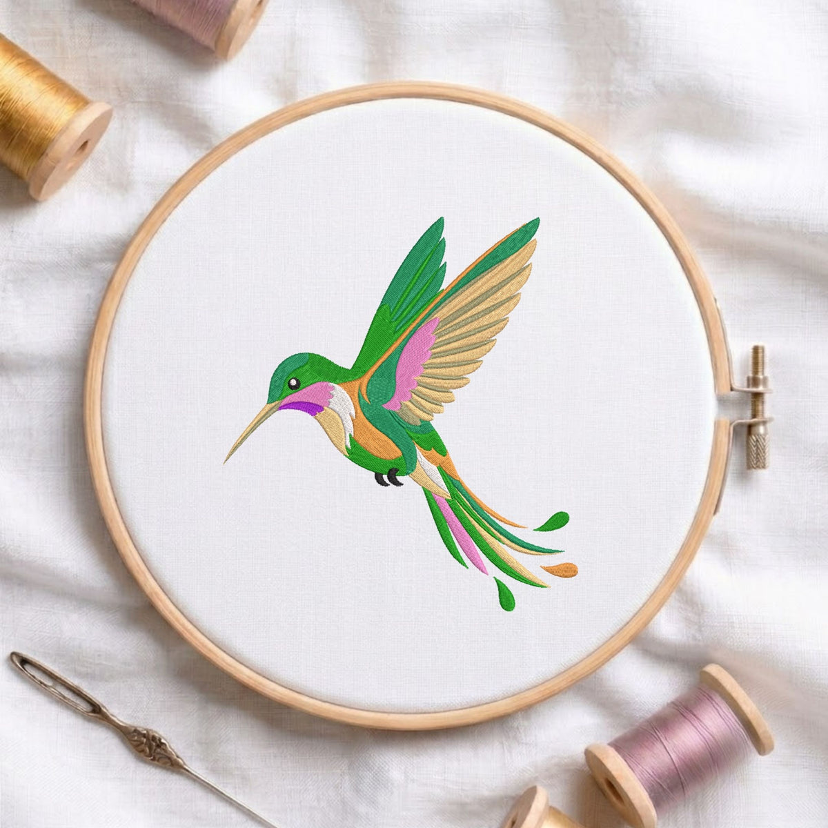 A circular embroidery hoop holding white fabric with a colorful hummingbird design in the center. The hoop is styled on a white fabric background with spools of thread and a needle nearby.