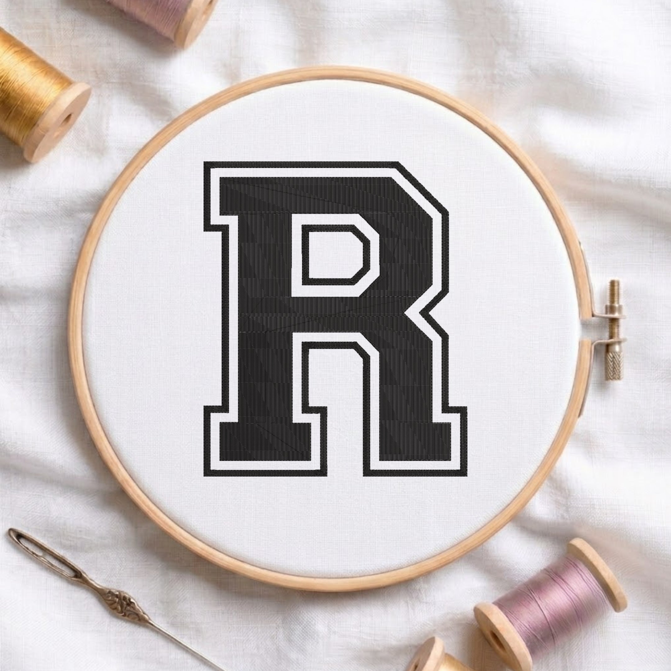Close-up view of an embroidered varsity-style block letter 'R' in black with a white and black outline, centered in a wooden embroidery hoop surrounded by spools of thread and a craft needle.