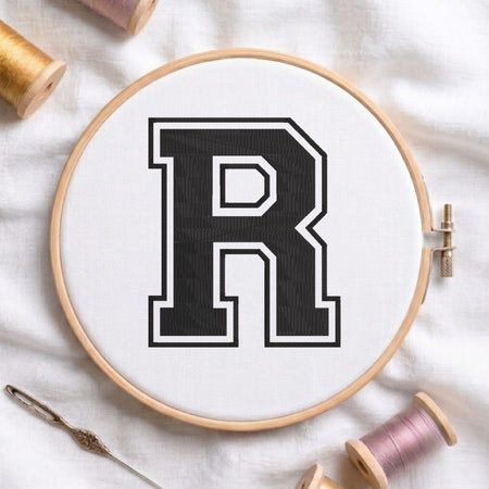 Close-up view of an embroidered varsity-style block letter 'R' in black with a white and black outline, centered in a wooden embroidery hoop surrounded by spools of thread and a craft needle.