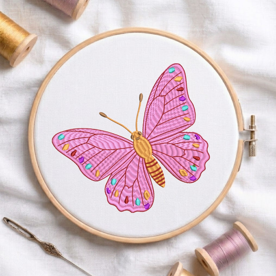 The pink butterfly embroidery design is secured in a wooden embroidery hoop, displayed alongside embroidery thread spools and a needle on a white fabric background.