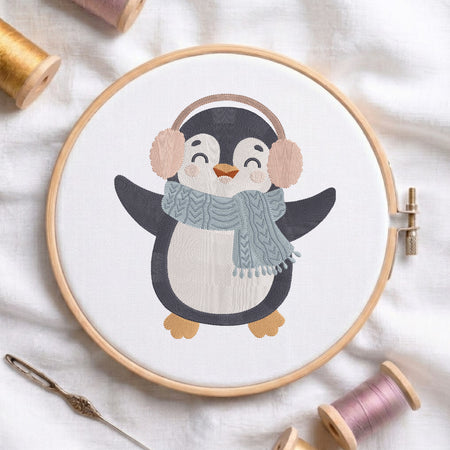A penguin embroidery design stitched inside a wooden embroidery hoop, surrounded by spools of thread and a needle on a soft white fabric background.