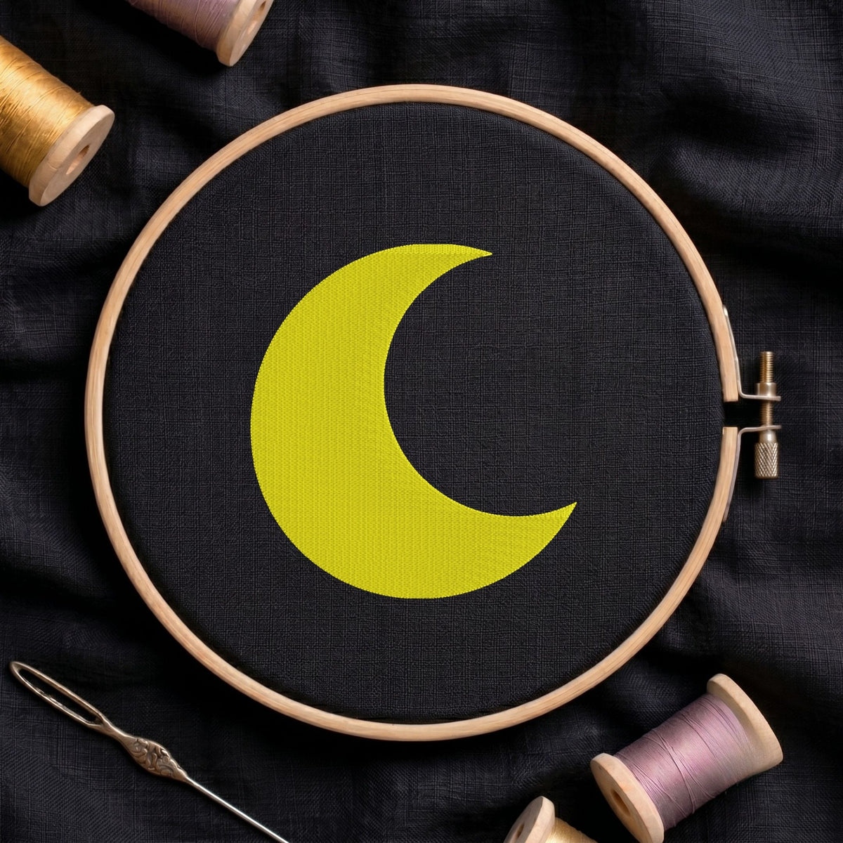 Bright yellow crescent moon machine embroidery design displayed inside a round wooden hoop on black fabric alongside thread spools and a needle.