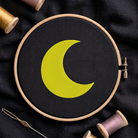 Bright yellow crescent moon machine embroidery design displayed inside a round wooden hoop on black fabric alongside thread spools and a needle.