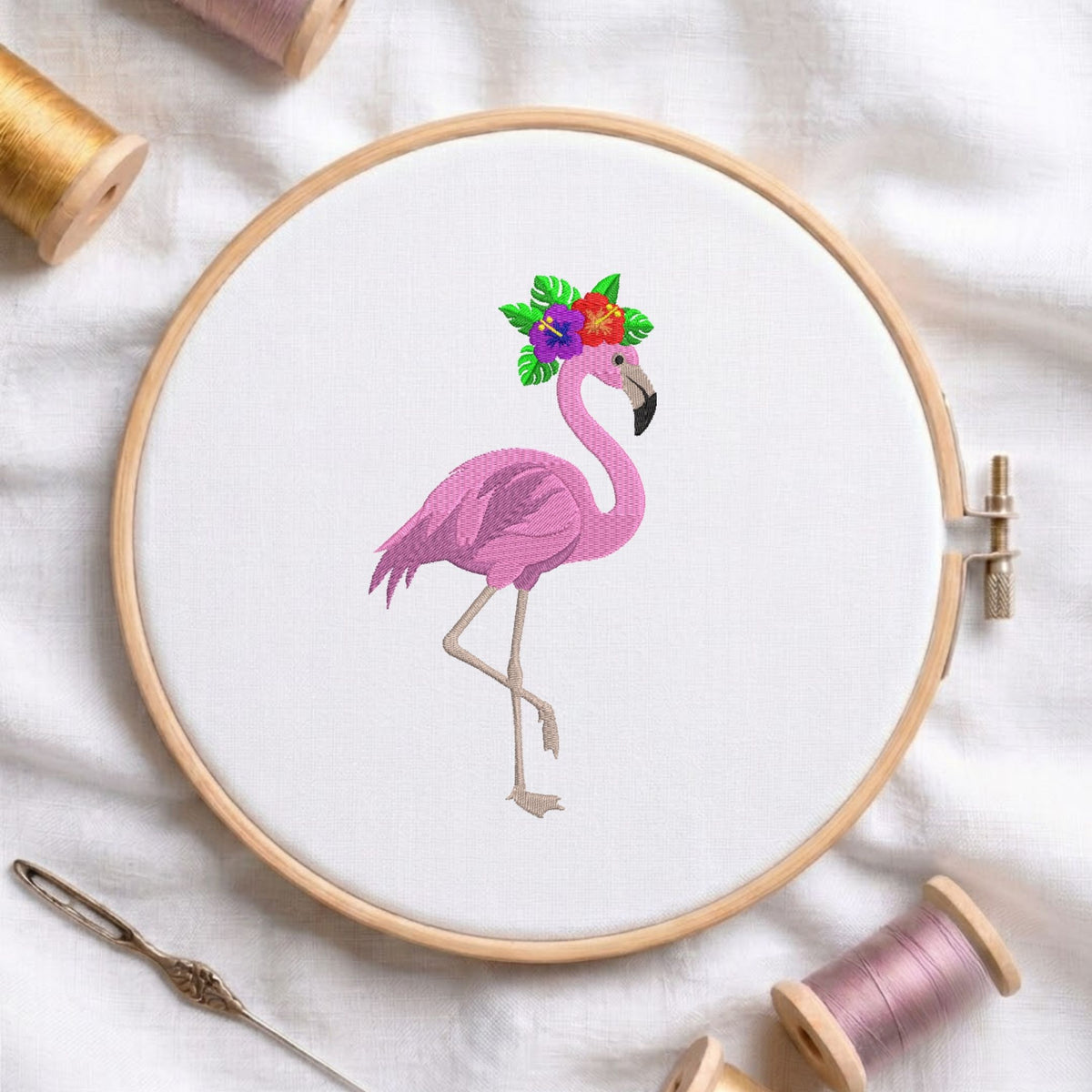 A circular embroidery hoop holding white fabric with a pink flamingo design wearing a colorful floral crown. The hoop is styled on a white fabric background with spools of thread, a needle, and wooden embroidery hoops nearby.
