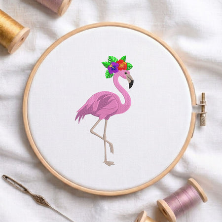 A circular embroidery hoop holding white fabric with a pink flamingo design wearing a colorful floral crown. The hoop is styled on a white fabric background with spools of thread, a needle, and wooden embroidery hoops nearby.