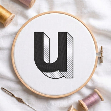 A wooden embroidery hoop holds white fabric with a large black letter 'U' outlined in black and filled with white and gray diagonal hatching. Spools of thread and a needle are arranged around it on a white fabric background.