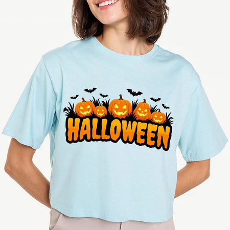 Woman wearing a light blue cropped t-shirt featuring an orange "HALLOWEEN" text design with glowing carved pumpkins, grass silhouettes, and flying bats.