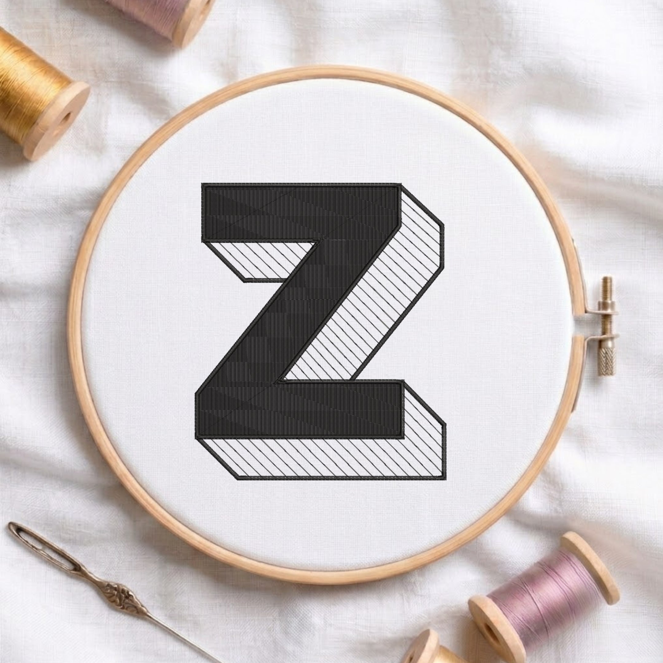 A wooden embroidery hoop holding white fabric with a large black letter Z, surrounded by spools of thread and a needle on a white fabric background.
