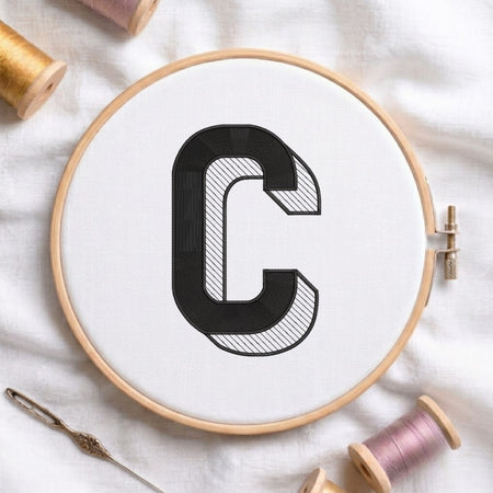 A wooden embroidery hoop holding white fabric with a large black letter C outlined and filled with hatched stitching. A needle lies at the lower left, and thread spools are visible around the hoop on a white fabric background.