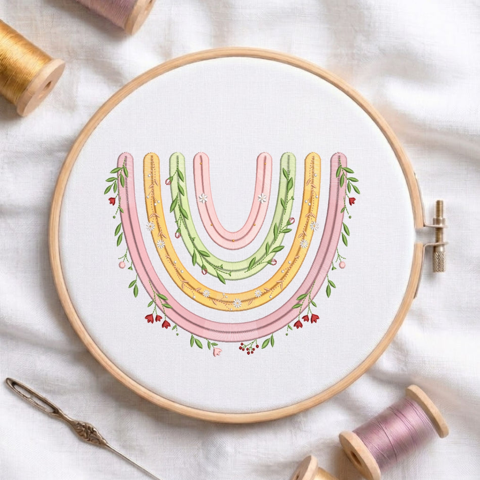 A circular embroidery hoop holding white fabric with a pastel rainbow design in pink, yellow, and green, surrounded by spools of thread and a needle on a white fabric background.