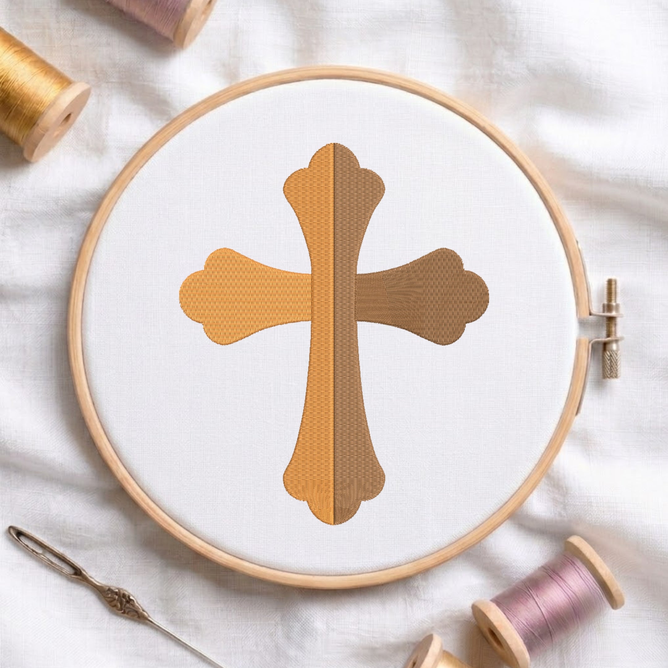 A circular embroidery hoop holding white fabric with a stitched cross design in warm gold and brown thread. The hoop is styled on a white fabric background with spools of thread and a needle around it.